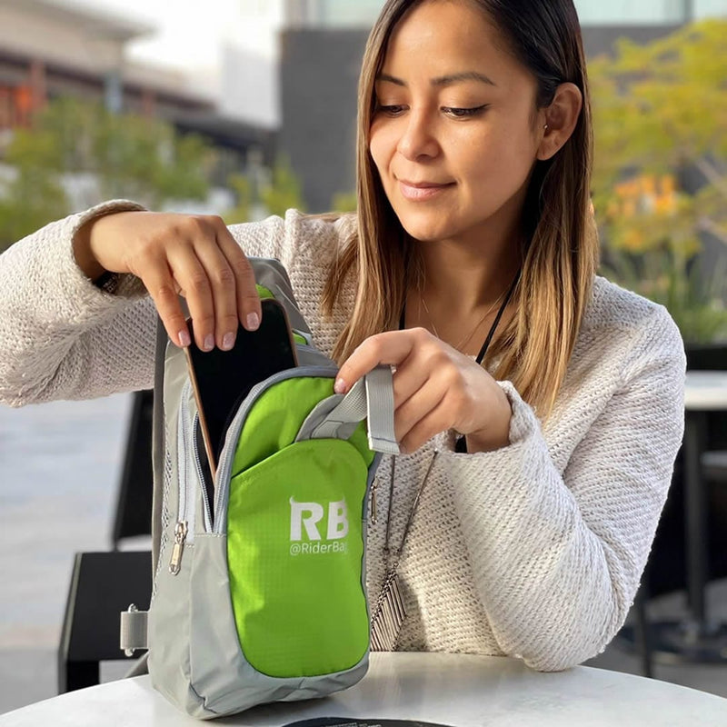 Laden Sie das Bild in Galerie -Viewer, Woman at an outdoor cafe table reaching into the Riderbag hi-vis green crossbody sling bag worn at her front